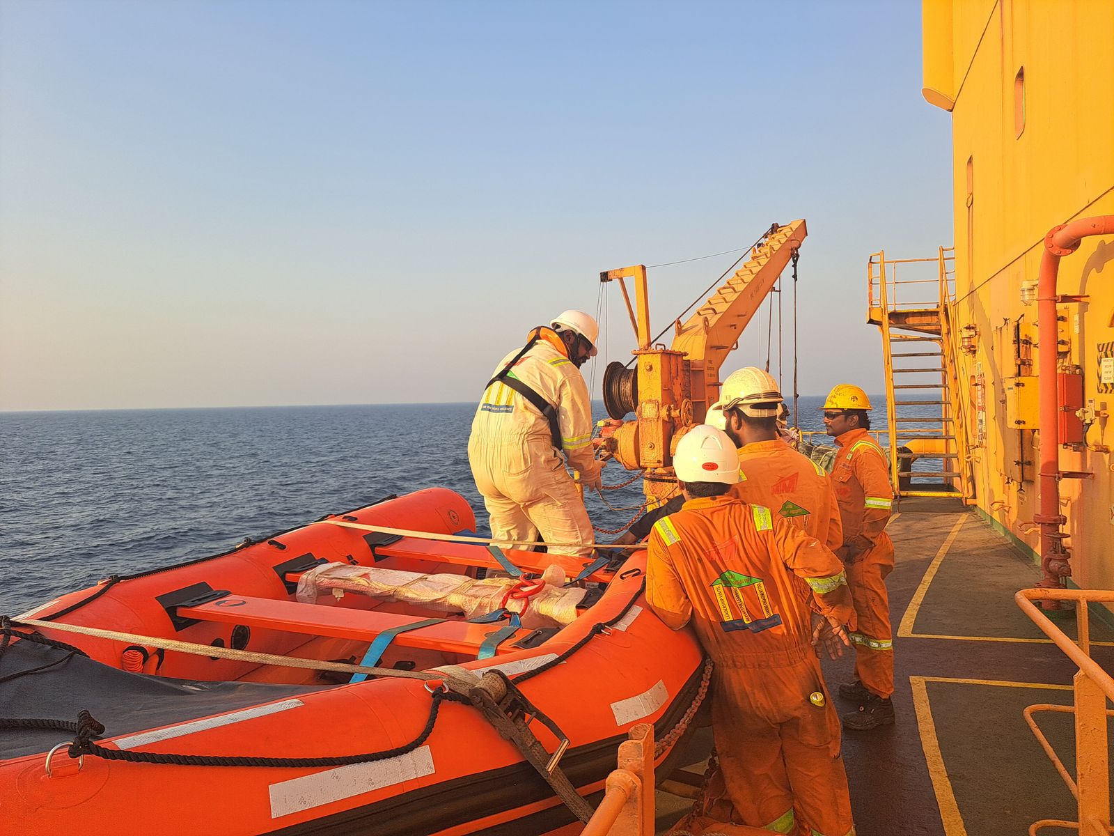 Offshore crew in coveralls and helmets working beside rescue craft on deck at golden hour